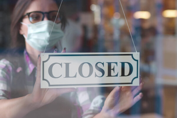 Through glass view of female business owner in safety mask hanging up sign with inscription closed on cafe door. Waitress in protective mask closing restaurant because of pandemic